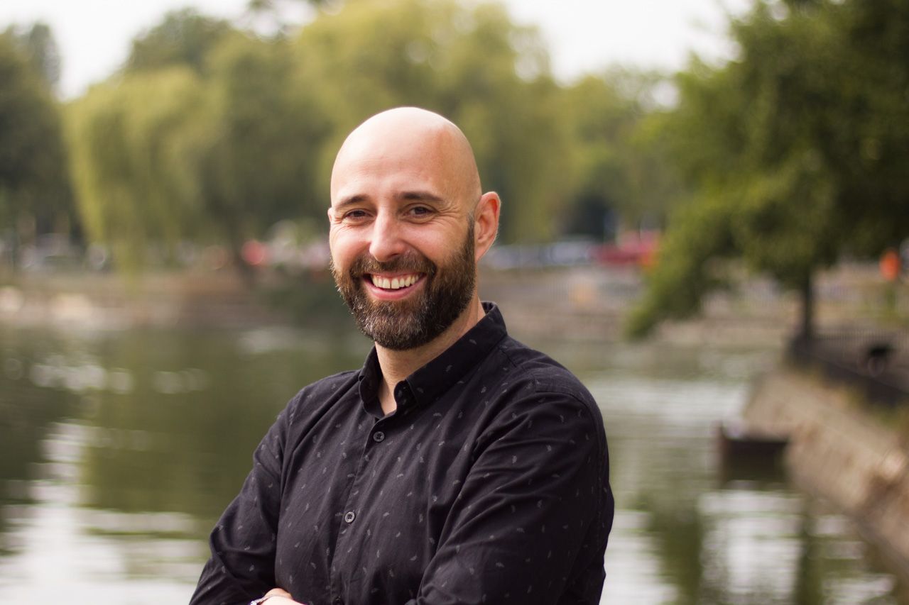 Man wearing a black shirt smiling at the camera with his arms crossed in front of a pond in a park