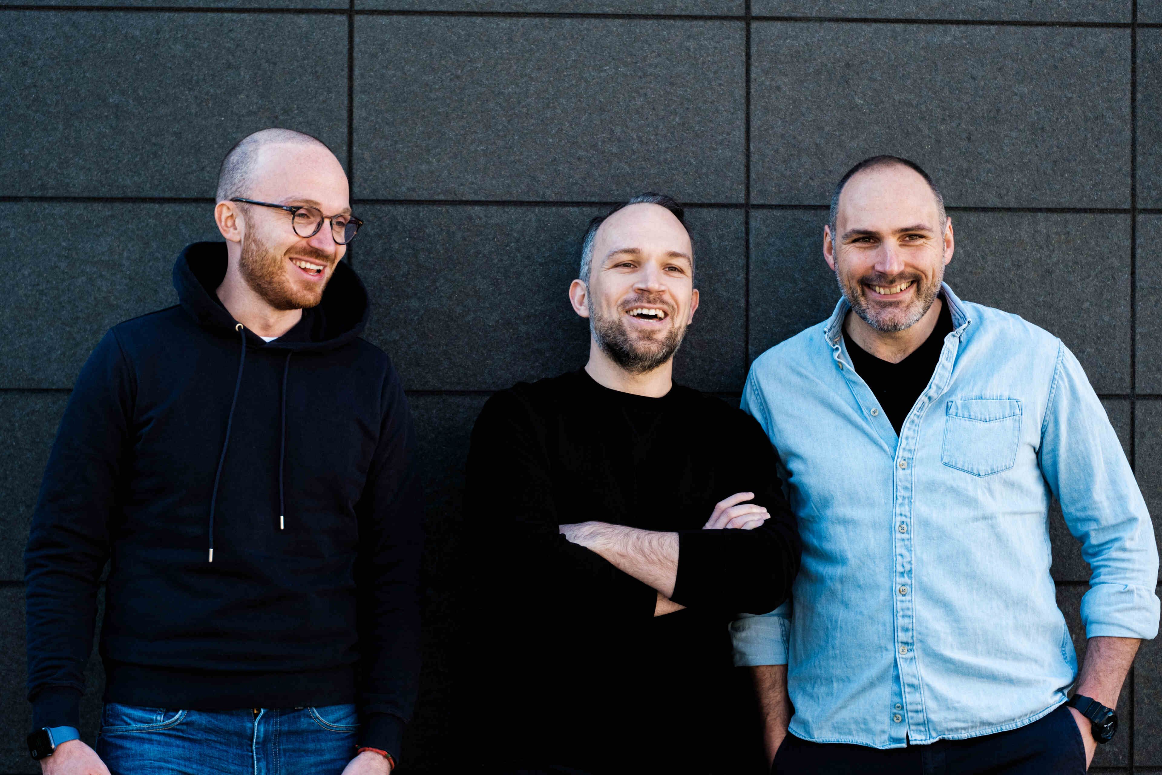Three smiling men posing in front of a grey acoustic wall