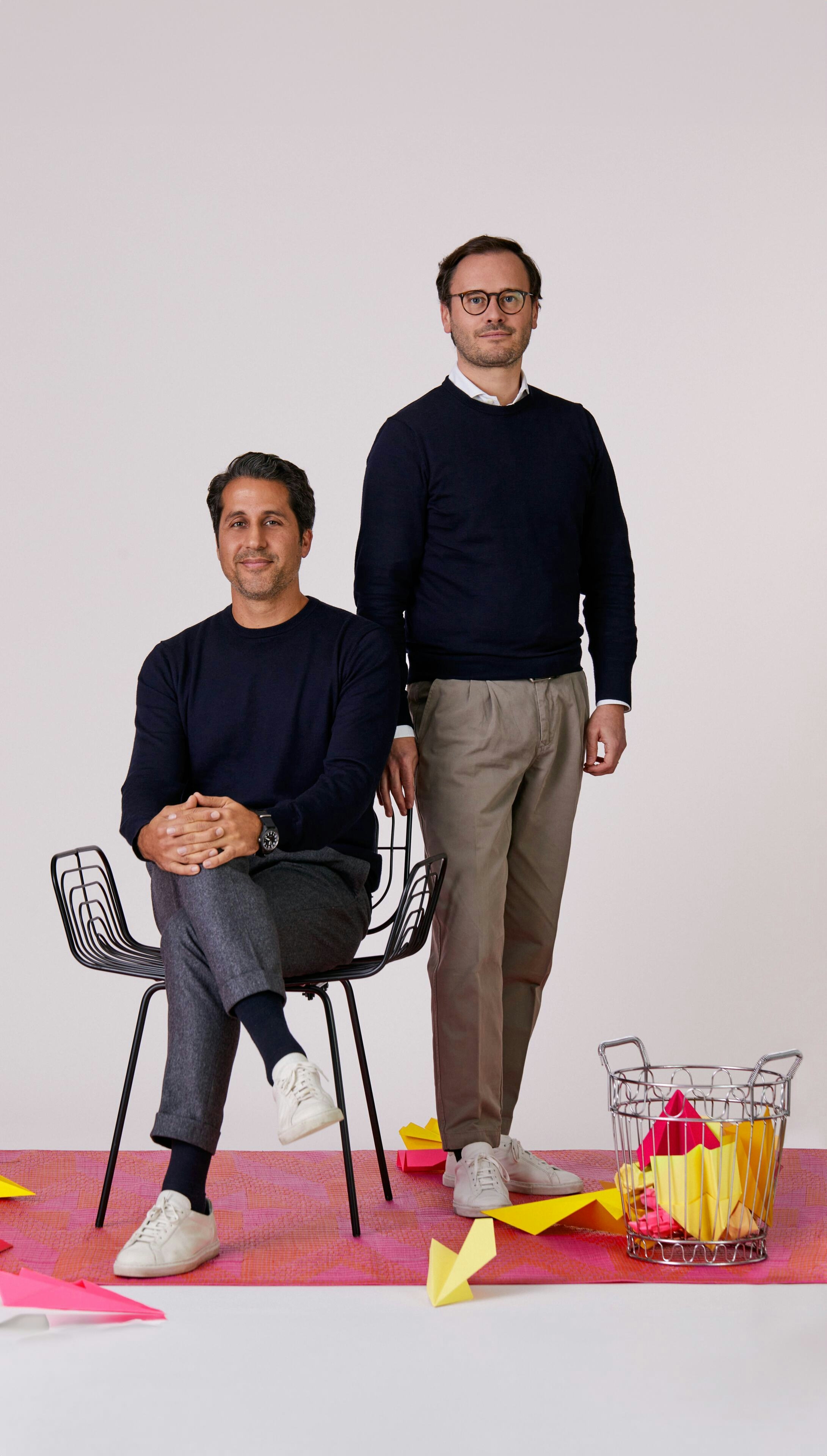 Two men posed in front of a grey background, one standing and one seated